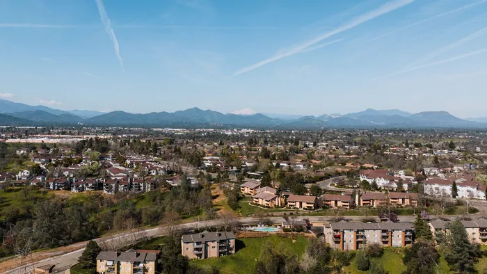 Homes and apartment complexes spread across a hillside in Redding, California, with forested mountains and a snow-capped peak on the horizon