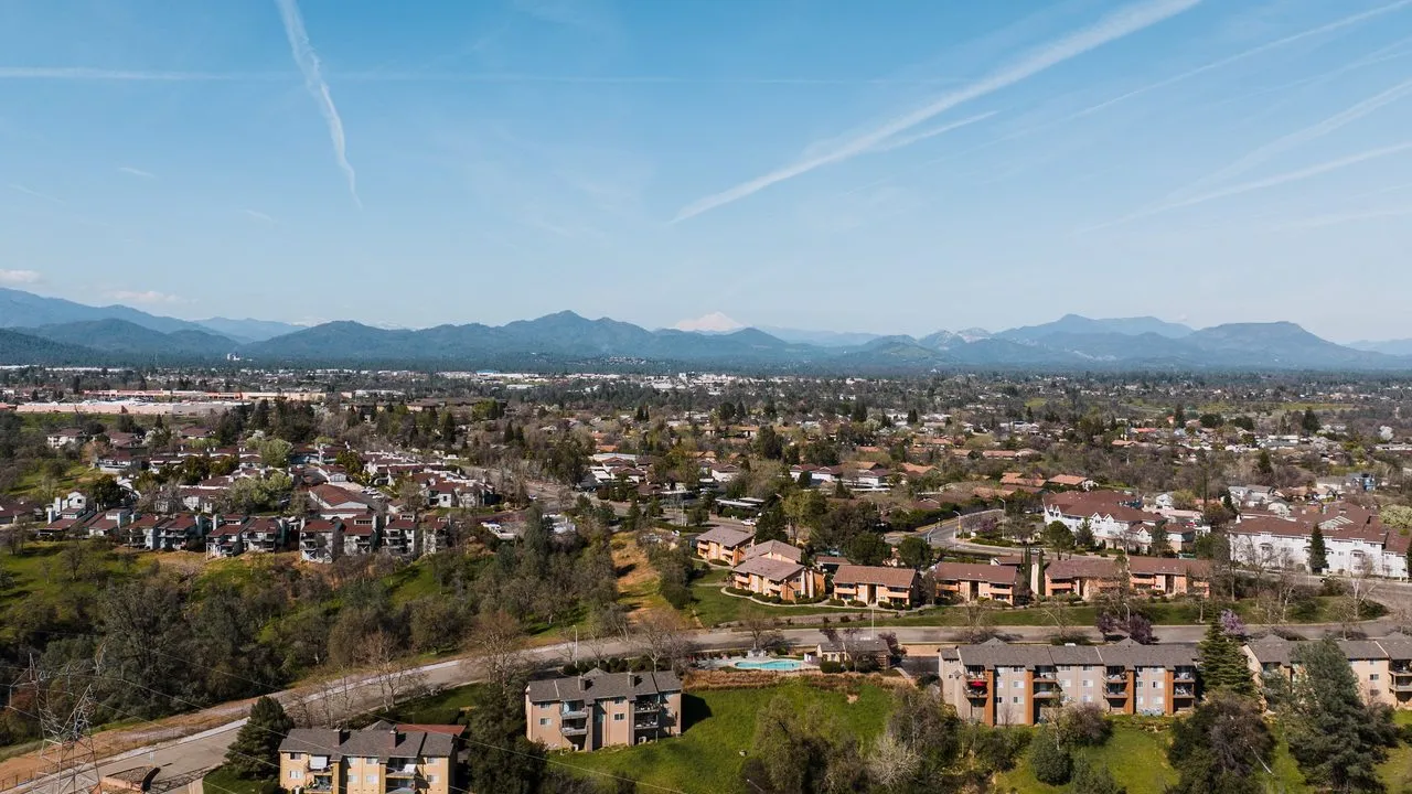 Homes and apartment complexes spread across a hillside in Redding, California, with forested mountains and a snow-capped peak on the horizon